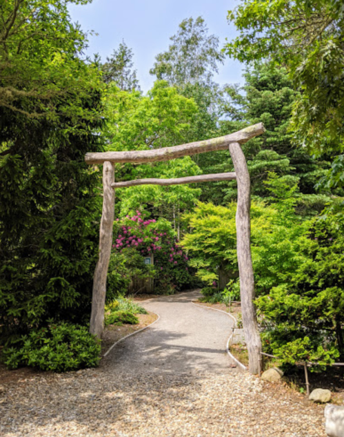 A wooden torii gate stands at the entrance of a lush, green garden path surrounded by trees and colorful flowers.