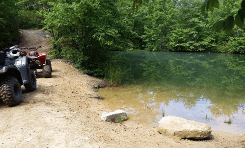 A dirt path beside a calm pond, with two ATVs parked nearby and lush greenery surrounding the area.