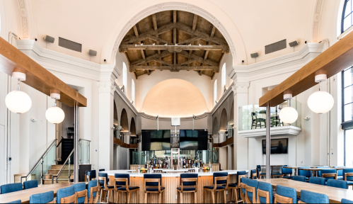 A spacious, modern restaurant interior featuring a high ceiling, wooden beams, and a stylish bar area with seating.