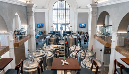 A spacious brewery interior with large metal tanks, high ceilings, and a bright entrance, featuring tables and patrons.