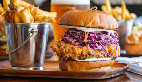 A crispy chicken sandwich with coleslaw and pickles, served with a side of fries in a metal bucket.