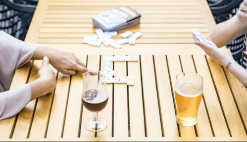 Two people playing dominoes on a wooden table, with drinks and a deck of cards nearby.