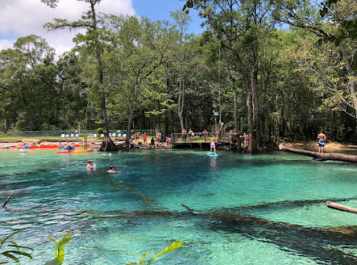 A serene natural swimming area with clear blue water, surrounded by trees and people enjoying various activities.