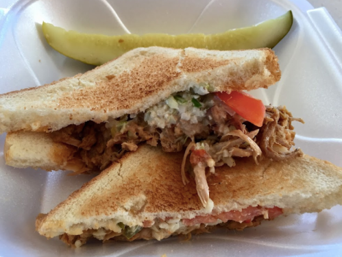 A toasted sandwich filled with shredded meat, tomatoes, and a creamy spread, served with a pickle on the side.