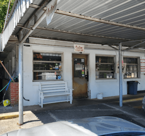 A small, weathered building with a covered entrance, featuring a bench and various signs on the walls.