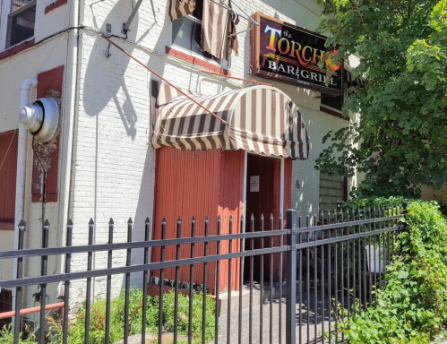 Exterior of The Torch Bar & Grill, featuring a striped awning and a fenced entrance on a sunny day.