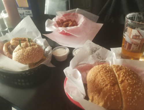 A table with two sesame seed burgers, a basket of fried snacks, dipping sauces, and a glass of beer.
