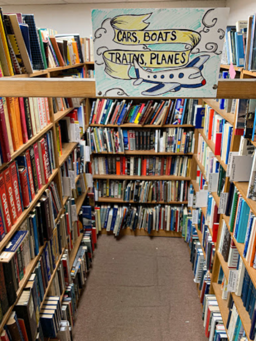 A narrow aisle between bookshelves filled with books, topped by a sign reading "Cars, Boats, Trains, Planes."