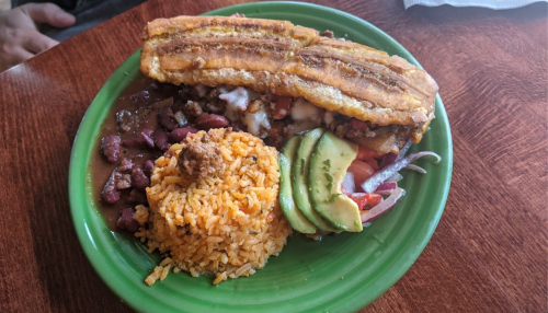 A vibrant plate featuring a sandwich with fried plantains, rice, beans, and fresh avocado slices.