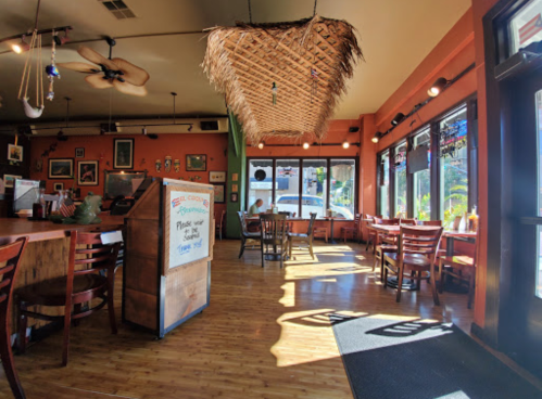 Cozy restaurant interior with wooden tables, a thatched ceiling, and sunlight streaming through large windows.