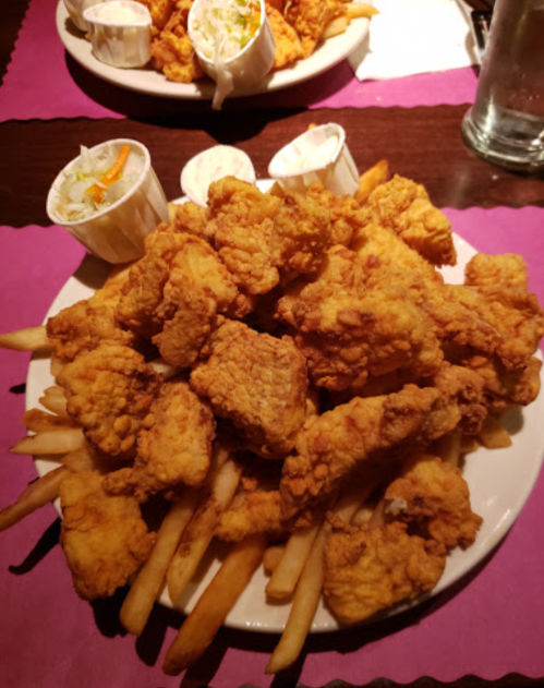 A large plate of crispy fried chicken pieces served over a bed of French fries, with two cups of coleslaw on the side.