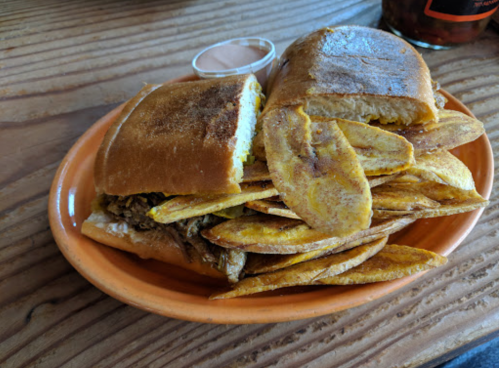 A plate with a sandwich and a generous serving of plantain chips, accompanied by a small dipping sauce.