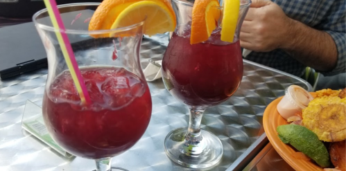 Two colorful drinks with orange slices on the rim, served in tall glasses, alongside a plate of food.