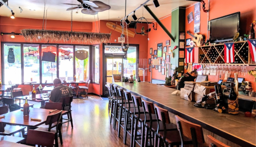 A vibrant restaurant interior with a long bar, tables, and large windows, featuring tropical decor and Puerto Rican flags.