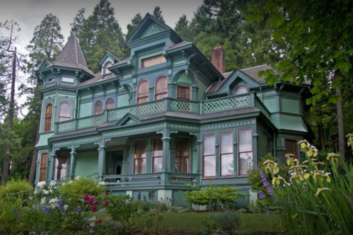 A large, ornate green Victorian house surrounded by lush greenery and colorful flowers.