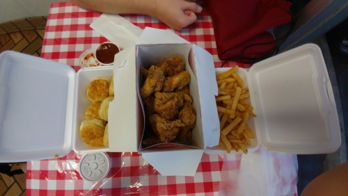A takeout box with fried chicken, biscuits, and fries on a red and white checkered tablecloth.