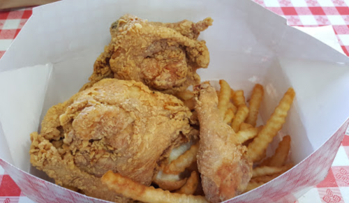 A takeout box containing crispy fried chicken pieces and crinkle-cut fries on a checkered tablecloth.