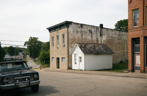 A weathered stone building and a small white house on a quiet street, with a vintage truck parked nearby.