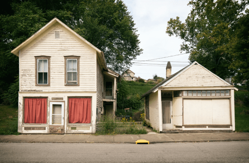 A weathered two-story house with red curtains and a small garage beside it, set against a green hillside.