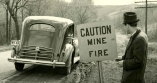 A vintage car parked near a sign warning of a mine fire, with a man in a hat observing the scene.