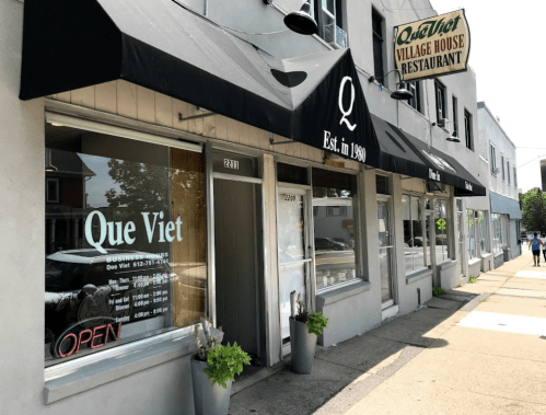 Exterior of Que Viet Village House Restaurant, featuring large windows, awnings, and an "OPEN" sign.