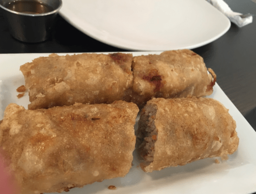 Three crispy spring rolls on a white plate, with one partially opened to reveal the filling. A small dipping sauce is nearby.