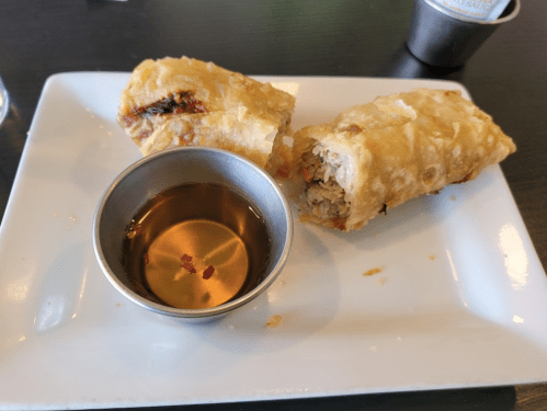 Two crispy spring rolls on a plate with a small bowl of dipping sauce.