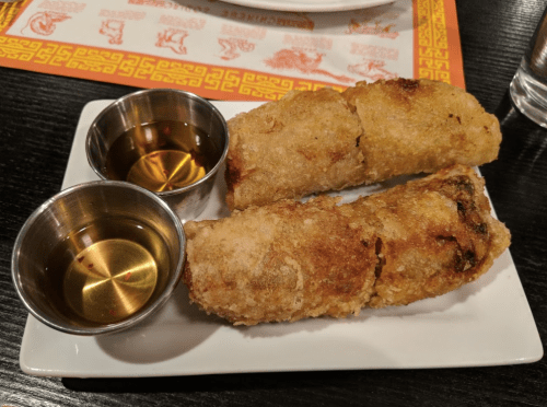 Two crispy spring rolls on a plate, served with two small bowls of dipping sauce.