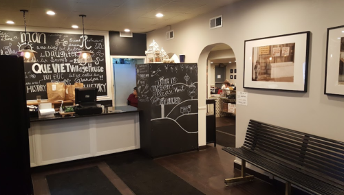 Interior of a cozy café with a chalkboard wall, a counter, and a bench, featuring framed photos and warm lighting.