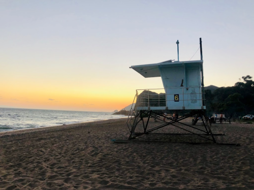 A lifeguard tower on a sandy beach at sunset, with calm waves and distant hills in the background.