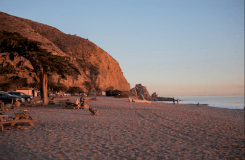 A serene beach at sunset, with cliffs in the background, picnic tables, and people walking along the shore.