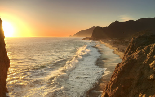 Sunset over a beach with gentle waves, rocky cliffs, and distant mountains, creating a serene coastal landscape.