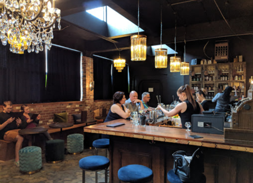 A stylish bar with a chandelier, patrons seated, and bartenders preparing drinks in a cozy, dimly lit atmosphere.