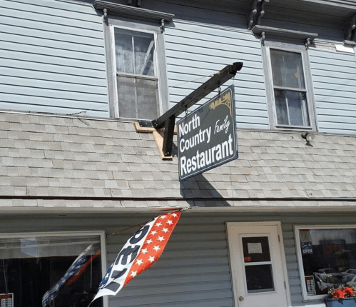 Sign for "North Country Restaurant" hanging above a building, with an open flag displayed in front.