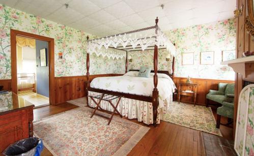 A cozy bedroom with a four-poster bed, floral wallpaper, wooden accents, and a small seating area.