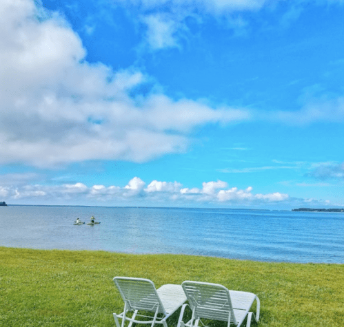 Two lounge chairs on green grass by a calm lake under a blue sky with fluffy clouds.