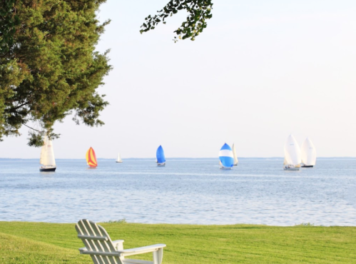 Sailboats with colorful sails on a calm lake, viewed from a grassy shore with a white chair in the foreground.