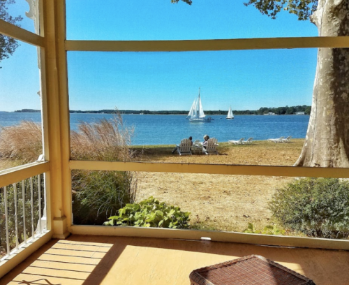 A serene view from a porch overlooking a lake with two sailboats and two people sitting on the shore.
