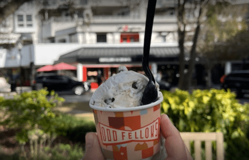A hand holding a cup of ice cream with chocolate chips, set against a blurred outdoor scene of a city street.