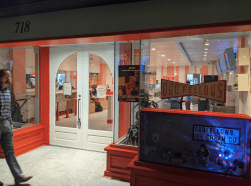 A storefront with a red and white theme, featuring "Odd Fellows Ice Cream Co." sign and a person walking by.
