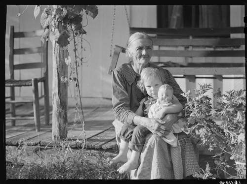 An elderly woman sits on a porch, holding a young child and a doll, with greenery and wooden chairs in the background.