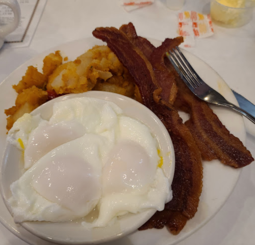 A plate with three poached eggs, crispy bacon, and hash browns, served with a fork on a white tablecloth.