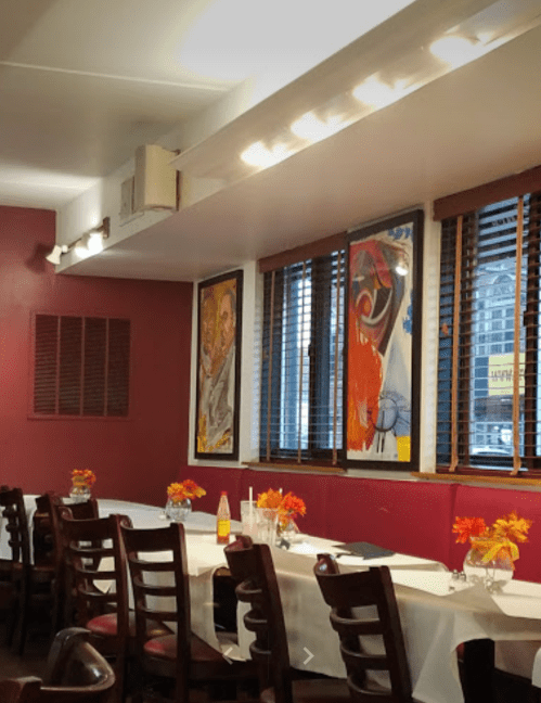 Interior of a restaurant with red walls, wooden chairs, and colorful artwork on the walls, featuring flower centerpieces.