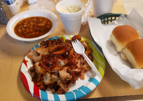 A colorful plate with BBQ meat, a side of baked beans, coleslaw, and two dinner rolls on a wooden table.