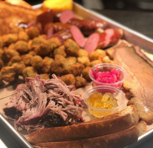 A platter of barbecue featuring pulled pork, brisket, sausage, fried okra, and colorful condiments on a tray.