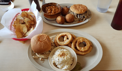 A table with two plates of food: pulled pork sandwiches, onion rings, coleslaw, baked beans, and potato chips.
