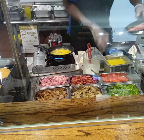 A food preparation station with various ingredients like meats, vegetables, and eggs being cooked on a stovetop.