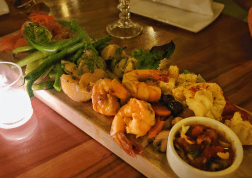 A wooden platter featuring assorted seafood, including shrimp and lobster, accompanied by a fresh vegetable garnish and dipping sauce.