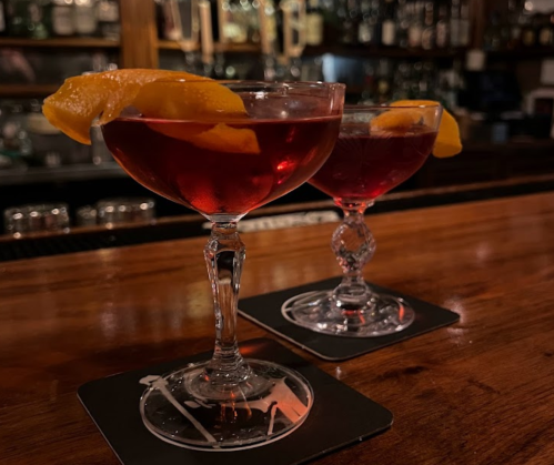 Two elegant cocktails with orange twists, sitting on a wooden bar top, with a blurred bar background.