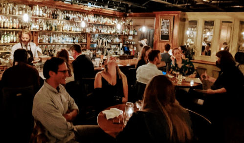 A lively bar scene with patrons enjoying drinks at tables, a bartender in the background, and warm ambient lighting.